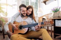 Cheerful tattooed male musician playing guitar near content female beloved while looking at each other in armchair in house room — Stock Photo