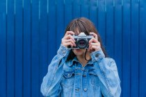 Anonymous female photographer in denim jacket taking picture on vintage photo camera on background of blue wall in city street — Stock Photo