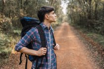 Explorador masculino con mochila caminando por sendero arenoso en el bosque durante el trekking y mirando hacia otro lado - foto de stock