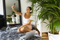 Очаровательная этническая женщина в беспроводной гарнитуре делает автопортрет на мобильном телефоне, сидя на ковре между зеркалом и ноутбуком дома — стоковое фото