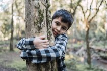 Дружелюбный этнический ребенок в клетчатой рубашке, обнимающий ствол дерева с мхом и лишайником, глядя в камеру в лесу — стоковое фото
