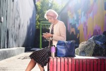 Вид сбоку триумфальной альтернативной женщины с окрашенными волосами, сидящей на скамейке в городском районе и просматривающей социальные сети на мобильном телефоне — стоковое фото