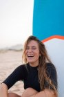 Cheerful female surfer sitting with blue SUP board on sandy seashore in summer and looking away — Stock Photo