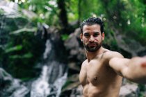 Conteúdo bonito macho com tronco nu tirando selfie e olhando para a câmera no fundo de rochas em bosques verdes — Fotografia de Stock