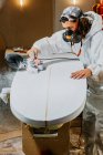 Male shaper in protective respirator and costume using electric planer and polishing surface of surfboard in workshop — Stock Photo