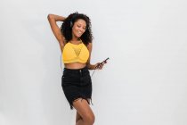 Mulher afro-americana feliz em fones de ouvido com celular dançando enquanto ouve música no fundo claro — Fotografia de Stock