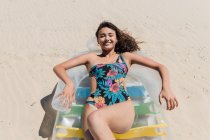 From above happy female in swimsuit lying on inflatable mattress on sandy seashore and sunbathing on sunny day during summer vacation — Stock Photo