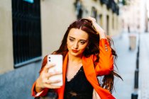 Stylish female with red hair and in vibrant orange jacket taking self shot on smartphone in city street — Stock Photo