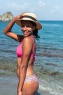 Вид збоку весела етнічна жінка-туристка в купальнику і капелюсі, дивлячись на камеру на узбережжі океану — стокове фото