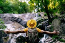 Обратный вид на неузнаваемого туриста-мужчину, стоящего на валуне и любующегося водопадом в лесу с распростертыми объятиями — стоковое фото