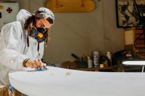 Seitenansicht eines männlichen Shaper in Sicherheitskostüm und Atemschutzmaske poliert Oberfläche des Surfbretts mit Handhobel in der Werkstatt — Stockfoto