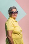 Felice donna moderna invecchiata con i capelli grigi e in occhiali da sole alla moda su sfondo rosa in studio e guardando la fotocamera — Foto stock