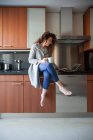 Geschäftsfrau mit lockigem Haar sitzt in der Küche und nimmt einen Aufguss, während sie ihren Laptop benutzt und zu Hause arbeitet — Stockfoto