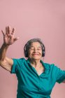 Gioiosa donna anziana moderna che ascolta musica in cuffia e balla a occhi chiusi su sfondo rosa in studio — Foto stock