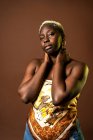 Modelo feminina afro-americana confiante vestindo cachecol de seda top tocando pescoço no fundo marrom em estúdio e olhando para a câmera — Fotografia de Stock
