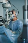 Médico adulto enfocado en máscara estéril y gorra médica ornamental mirando a través del microscopio quirúrgico contra compañero de trabajo en el hospital - foto de stock