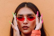 Портрет харизматичной женщины с длинными яркими ногтями, надевающей модные солнцезащитные очки на оранжевую стену, смотрящей в камеру — стоковое фото