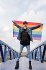 Von unten ein homosexueller Mann, der mit erhobener Regenbogenfahne auf einer Brücke steht und in die Kamera blickt — Stockfoto