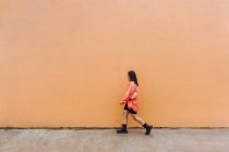 Ganzkörper-Seitenansicht einer stilvollen Frau in trendigem Outfit und Sonnenbrille, die an orangefarbener Wand vorbeiläuft — Stockfoto