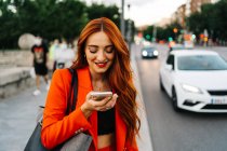 Smiling female with red hair and in orange suit recording audio message on mobile phone while communicating with friend on social media and walking in city street — Stock Photo