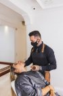 Homem em máscara manchando fundação de cara de mulher loira durante o trabalho em estúdio de maquiagem profissional — Fotografia de Stock