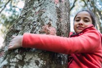 Знизу чарівна етнічна дитина торкається грубої кори старого стовбура дерева з лишайником, дивлячись на камеру в лісі — стокове фото