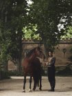 Piena lunghezza di femmina nera adulta in abito elegante in piedi con cavallo marrone vicino a alberi verdi e recinzione del castello di giorno in cortile — Foto stock