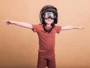 Веселый ребенок в шлеме и защитных очках смотрит в камеру с протянутыми руками на бежевом фоне — стоковое фото