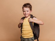 Positif cool écolier pré-adolescente avec sac à dos regardant la caméra sur fond brun en studio — Photo de stock
