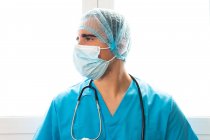 Вид збоку чоловічого лікаря в масці і форма, що стоїть біля вікна в клініці — стокове фото