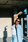 Vue latérale de la femme ethnique excitée en tenue élégante et lunettes de soleil s'amusant sur le trottoir urbain à la lumière du soleil — Photo de stock