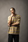 Young thoughtful African American male model with braided hair dressed in oversized striped shirt and necklace looking at camera against gray background — Stock Photo