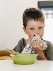 Lindo niño limpiando la boca con servilleta mientras está sentado en la mesa con un tazón de sopa de crema durante el almuerzo en casa - foto de stock