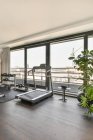 Interior of spacious hall with treadmill and sports supplies in front of large panoramic window overlooking cityscape in modern luxurious apartment — Stock Photo