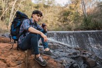 Caminhante masculino sedento com mochila bebendo água do copo com palha enquanto sentado na rocha perto de cachoeira na floresta e olhar para longe durante o intervalo — Fotografia de Stock