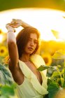 Gracieuse jeune femme hispanique en robe jaune élégante debout avec les bras levés au milieu de tournesols en fleurs dans le champ de campagne dans la journée ensoleillée d'été en regardant la caméra — Photo de stock