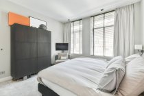 Інтер'єр сучасної світлої спальні з м'яким ліжком і дерев'яним шафою розміщений на килимі біля вікна з шторами і телевізором в кутку — стокове фото