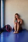 Весела жінка-спортсменка сидить на килимку зі спортивною сумкою в спортзалі і переглядає мобільний телефон — стокове фото