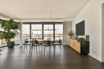 Dining table and chairs placed near large panoramic window in spacious room with TV and cabinet in modern loft style apartment — Stock Photo