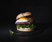 Hamburger savoureux avec oeuf posé sur galette et laitue fraîche servie sur planche d'ardoise sur fond noir en studio — Photo de stock