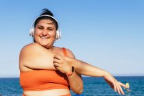 Cheerful ethnic female athlete with curvy body listening to song from headphones while looking at camera and does stretches under a blue sky — Stock Photo
