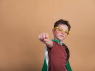 Kind in Superhelden-Umhang und dekorativer Brille zeigt vor Kamera Kraftgeste — Stockfoto