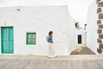 Vue latérale de la femelle avec sac à dos marchant sur la chaussée contre les maisons blanches et ciel gris nuageux sur la rue de la ville à Fuerteventura, Espagne — Photo de stock