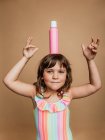 Cheerful preteen girl in swimsuit standing with bottle of sunblock cream on head on brown background in studio and looking at camera — Stock Photo