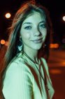 Вид сбоку очаровательной молодой женщины с длинными волосами, смотрящей в камеру на проезжую часть в вечернем городе — стоковое фото