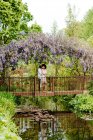 Vue à distance du couple aimant étreignant sur le pont sur l'étang tout en se tenant sous l'arc avec des fleurs de glycine en fleurs dans le jardin naturel — Photo de stock