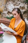 Serena tenera lettura femminile libro interessante mentre seduto a tavola sulla terrazza estiva della casa — Foto stock