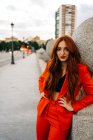 Mulher encantadora com cabelos vermelhos longos e em terno laranja na moda em pé na rua à noite e olhando para a câmera — Fotografia de Stock