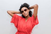 Jeune femme afro-américaine à la mode avec des cheveux bouclés en rouge et des lunettes de soleil regardant la caméra — Photo de stock