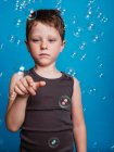 Astonished preteen boy showing touching gesture with index finger in air in studio with flying soap bubbles on blue background — Stock Photo